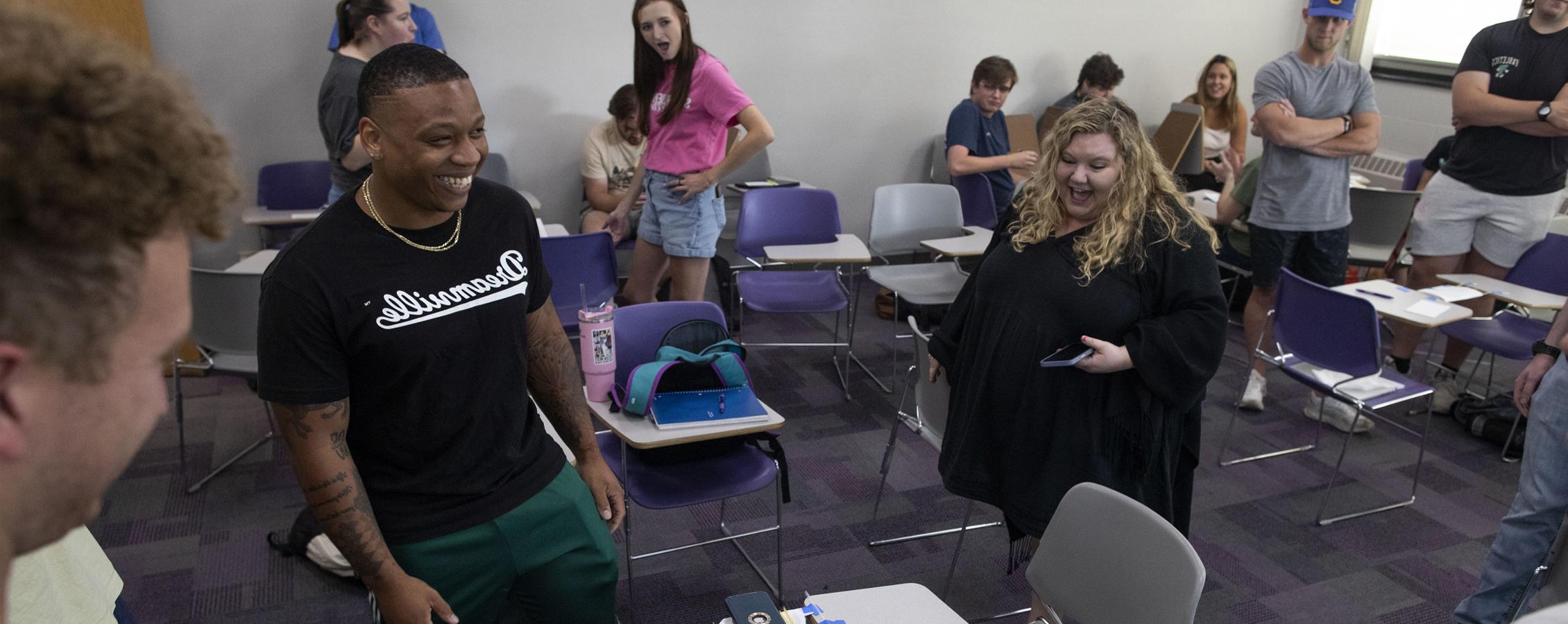 Students in a classroom cheer as one student smiles, standing amongst desks and chairs.