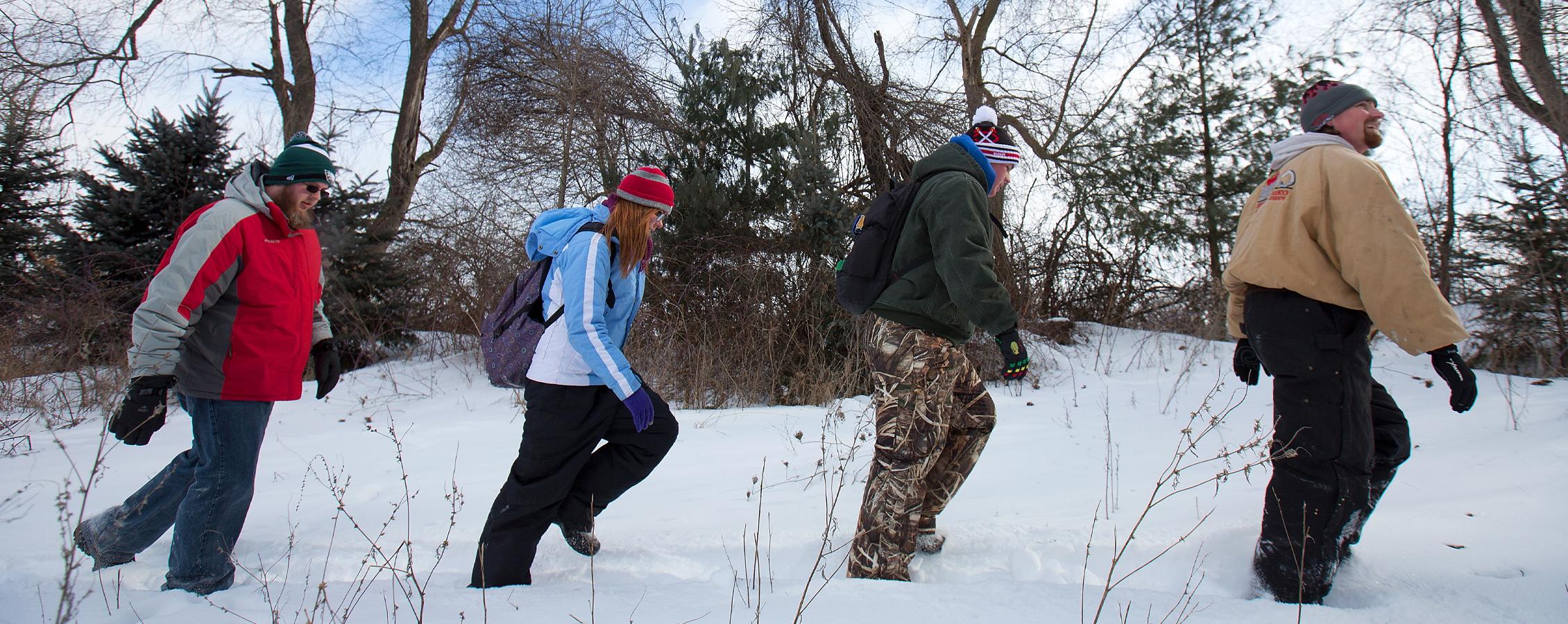 Four students walk through the snow in a wooded area.