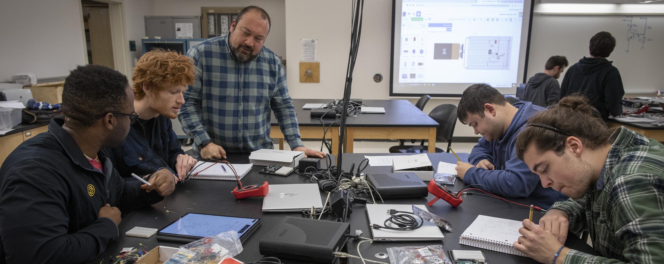 Students and a faculty member gather around a table as as they build circuitry. 