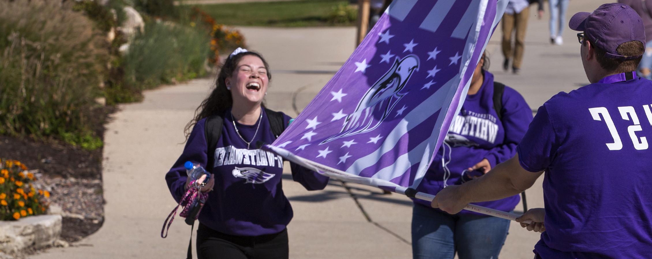 Three students laugh together, waving a purple Warhawk flag.