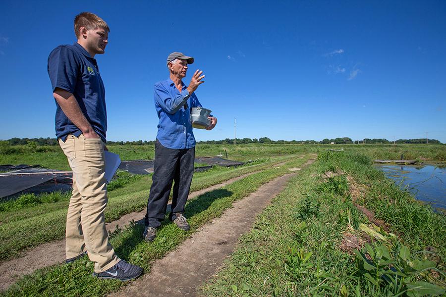 Two people stand in a field with a small body of water.