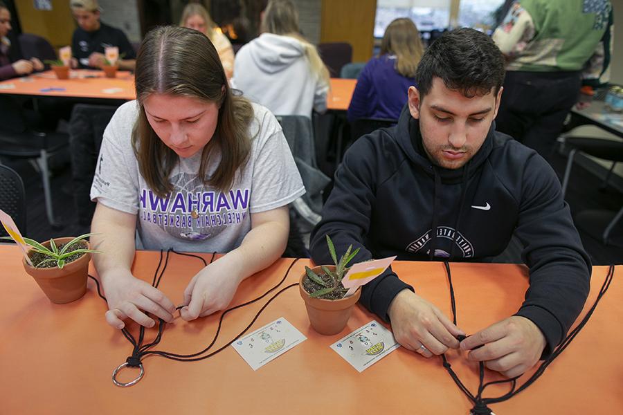 Two students sit at a table and assemble macrame plant hangers with succulents in small pots.