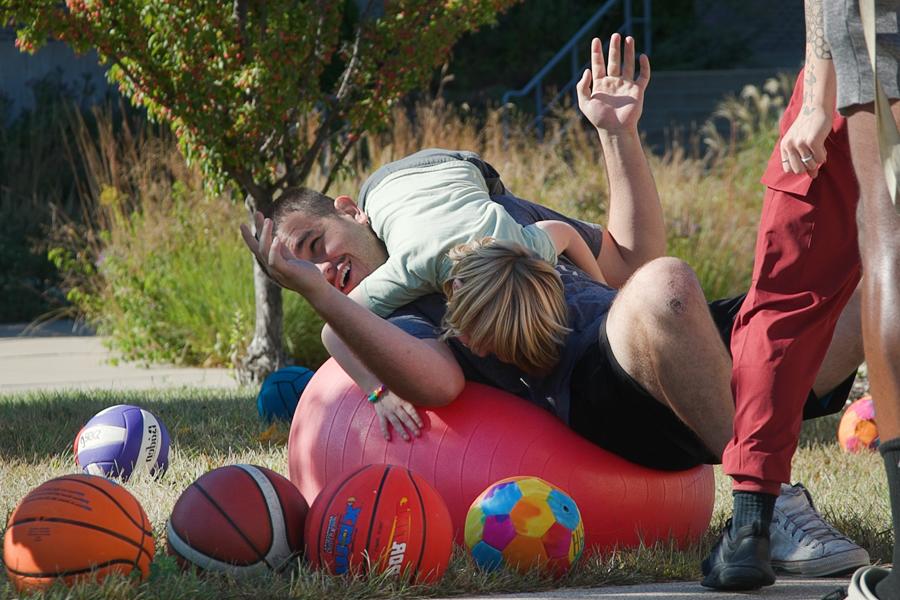 A student lays on a large red exercise ball and laughs as a child jumps on him.