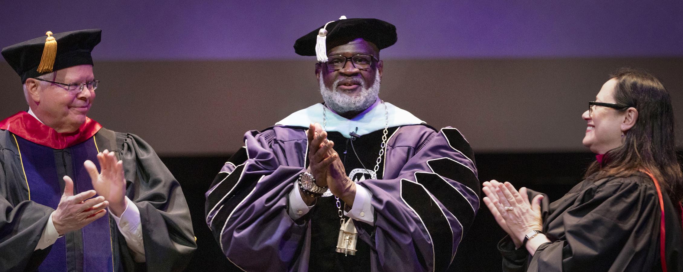 Chancellor King stands with two other people on stage, all dressed in academic regalia.