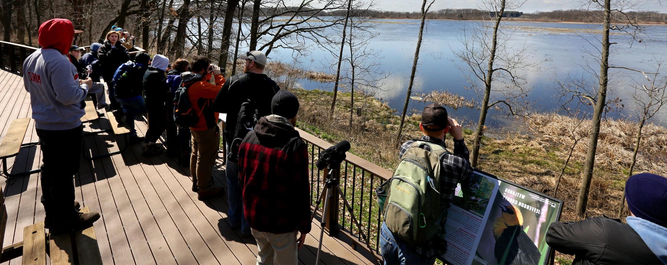 Students stand on a deck and look over Rose Lake State Natural Area through binoculars.