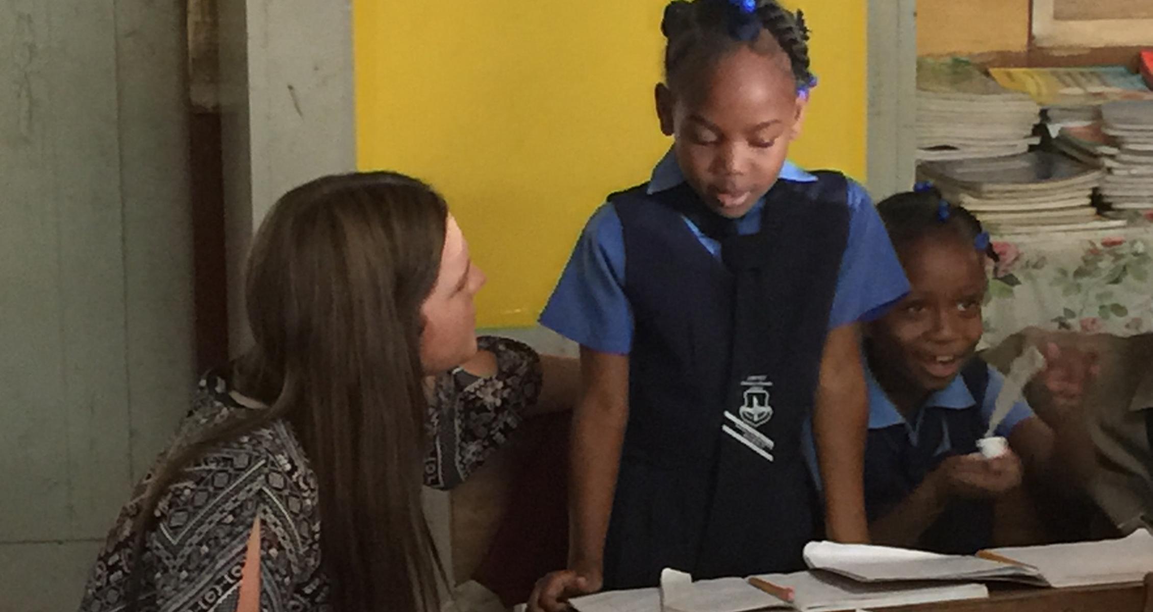 A 足彩平台 student kneels by two Jamacian girls in a Jamacian classroom.