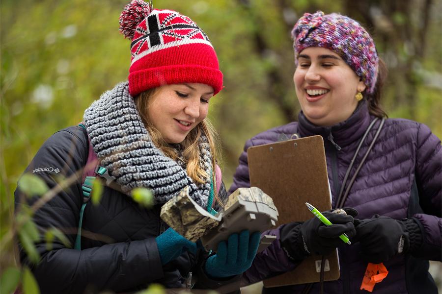 Two women are outside looking at a motion detecting trail camera.