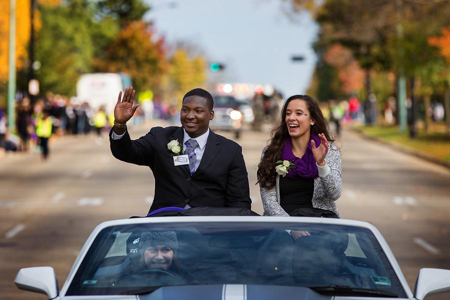 Two people wave from a convertible car during a Homecoming parade.